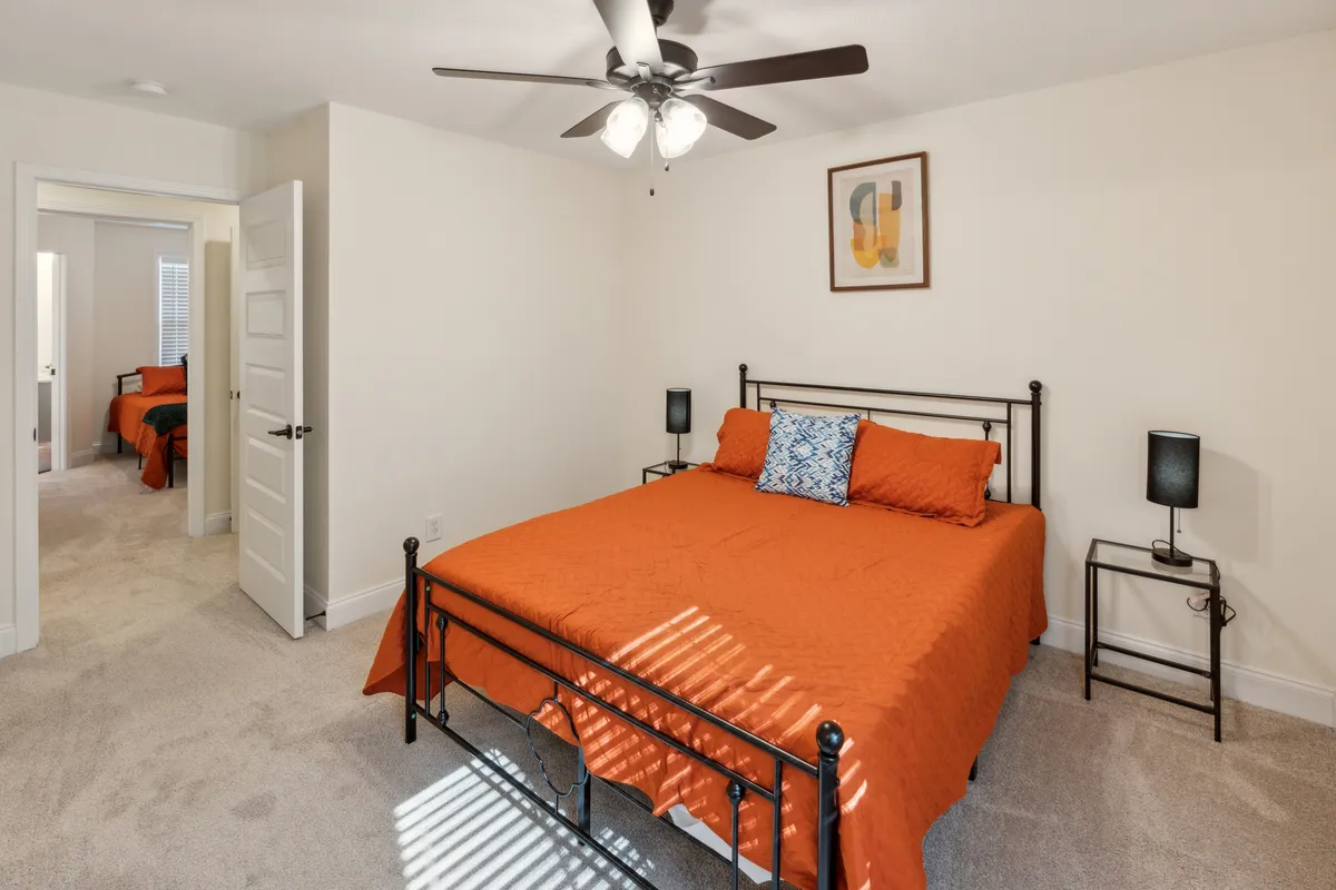 A cozy bedroom with an orange bedspread, contemporary wall art, a nightstand with a lamp, and an open door leading to another room. The arrangement conveys warmth and the possibility of both privacy and shared family moments, such as reading or winding down before bed.