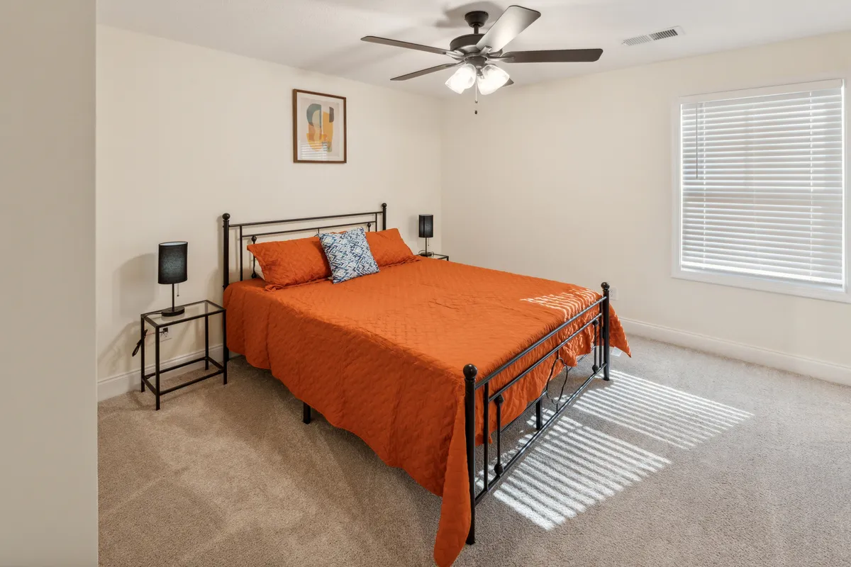 A well-appointed bedroom featuring an orange bedspread, decorative pillow, framed art above the bed, and matching nightstands with lamps. Natural light fills the space, creating a warm and inviting atmosphere ideal for relaxation.