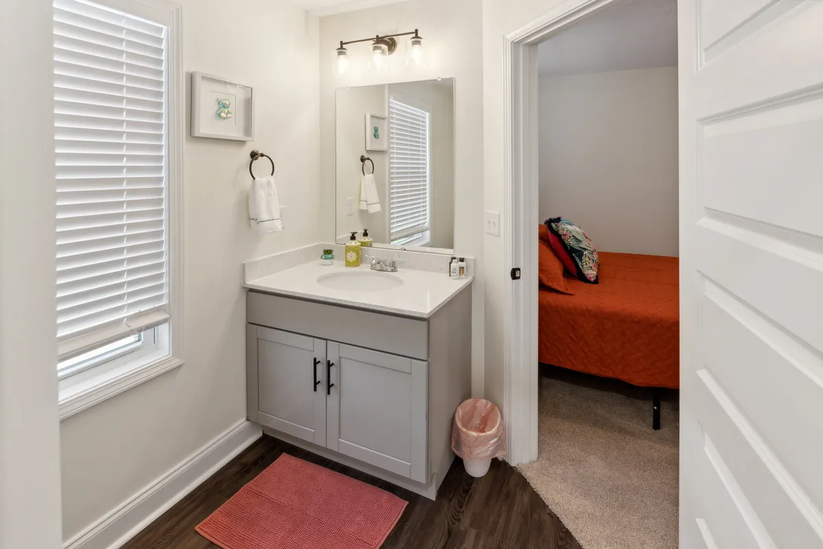 A bright and organized bathroom scene featuring a vanity with sink, a large mirror, tasteful decor, and a doorway leading toward the bedroom. The space is both practical and serene, offering a refreshing place for guests to unwind after a busy day out.