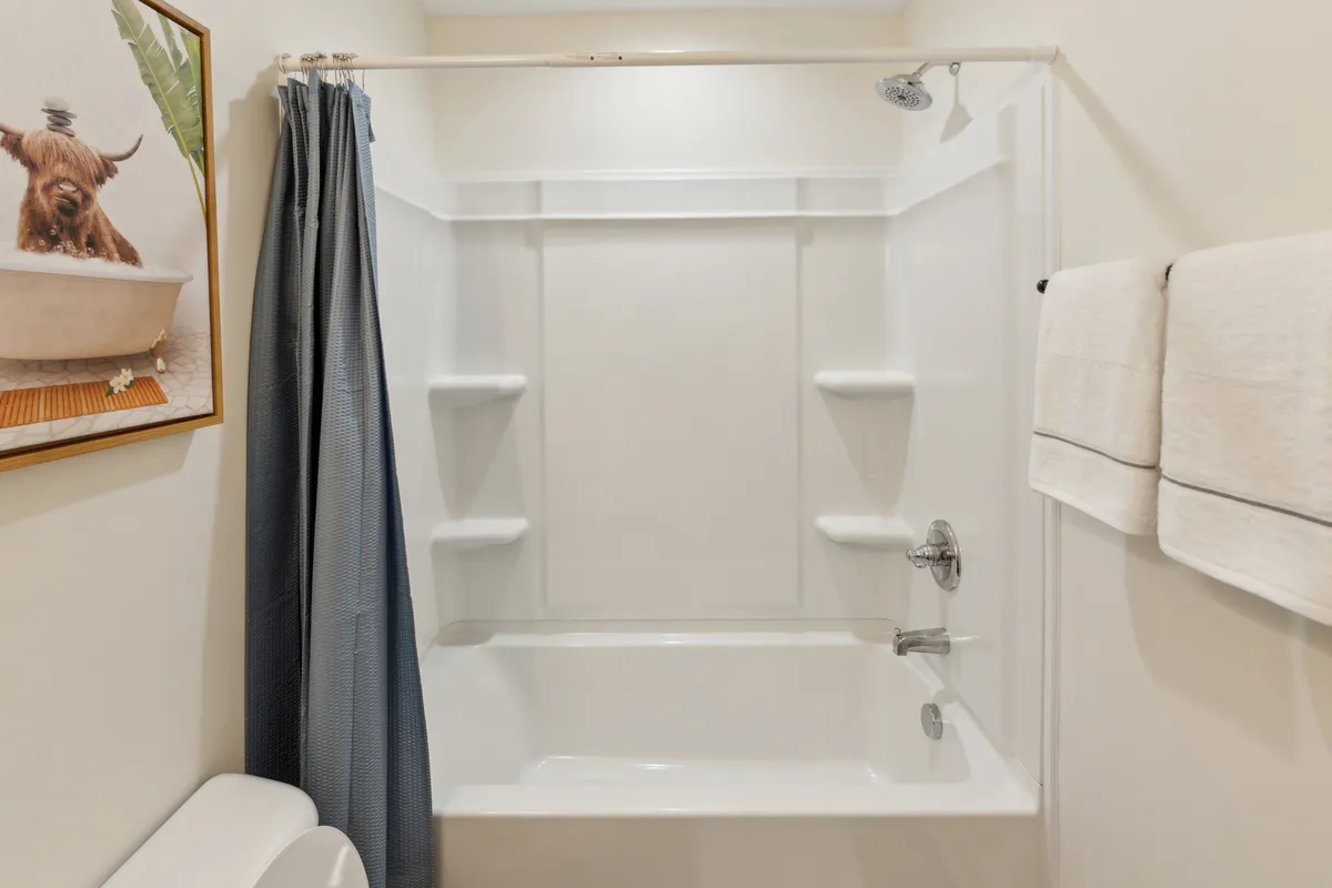 A calming bathroom with a shower-bath area, blue shower curtain, fluffy white towels, and unique dog-themed artwork on the wall. The space feels serene and is well-suited for self-care and relaxation.