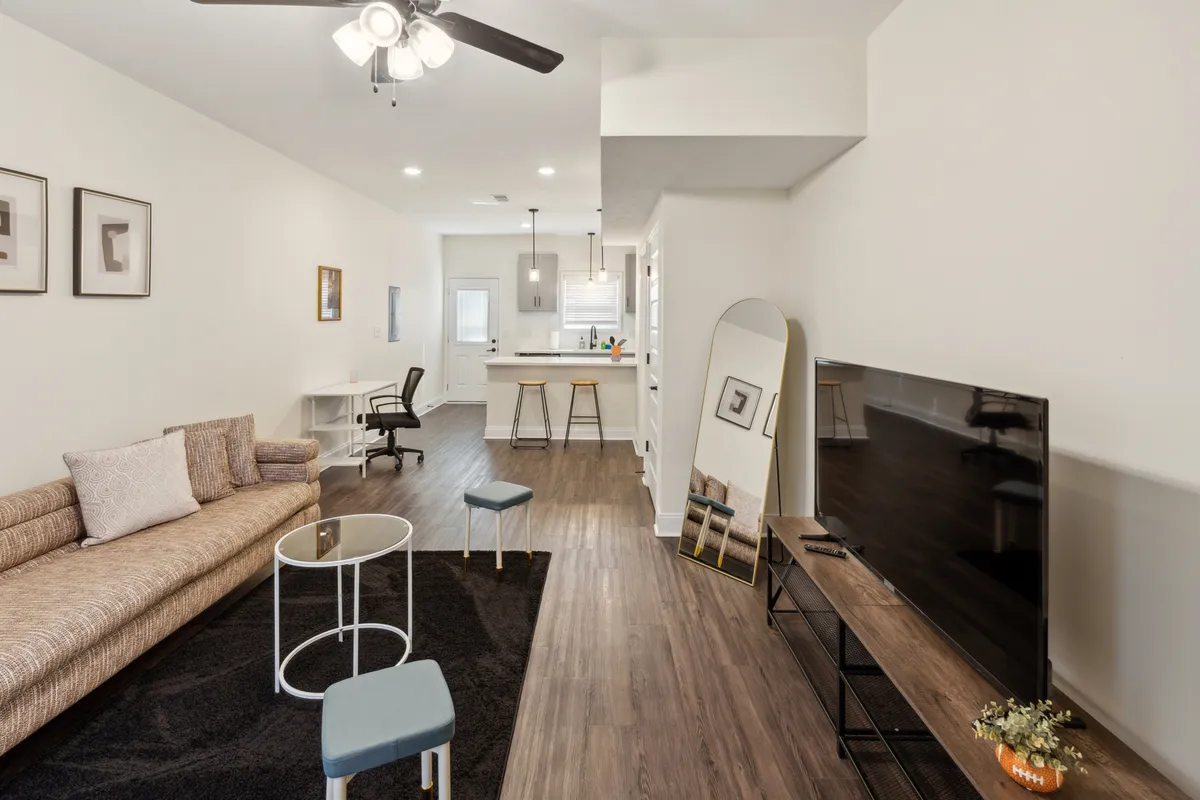 This interior shot captures a modern open-plan living space with a sofa, coffee table, television, a partial view of the kitchen area, and a prominent mirror. The layout is airy and contemporary, ideal for both relaxing and entertaining.